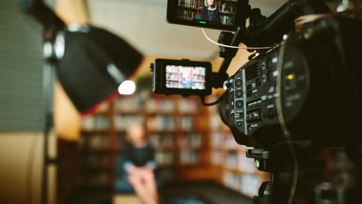 Out of focus, a person sits to the rear of a library. In the foreground, a video camera is visible to capture their interview.